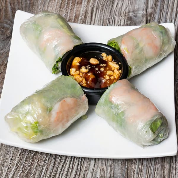 Four fresh spring rolls with shrimp and lettuce, arranged around a peanut dipping sauce on a white plate.
